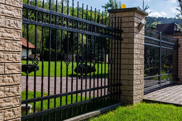 A black wrought iron fence with intricate details between stone pillars, set against a green lawn and paved driveway.