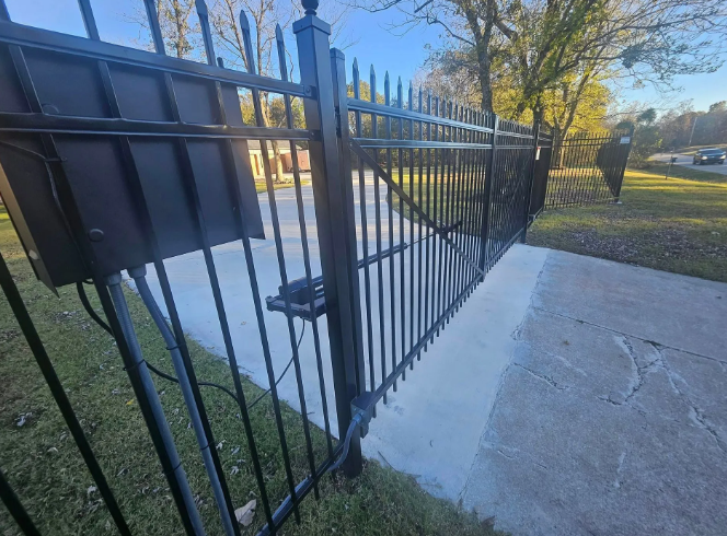 Black metal driveway gate with an electronic operator box and arm mounted on a post beside a concrete driveway.