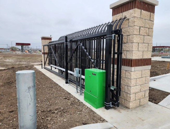 A black metal sliding gate with an arched top, powered by a green control box, set between two brick and stone pillars.
