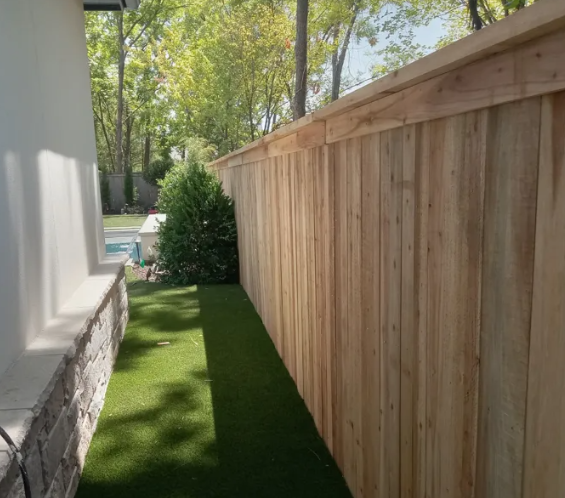 A narrow side yard with artificial turf, a light-colored stone-base wall on the left, and a tall cedar privacy fence.