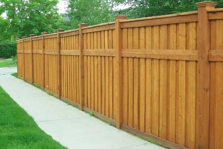 A tall, stained wooden privacy fence runs alongside a concrete sidewalk next to a grassy lawn.
