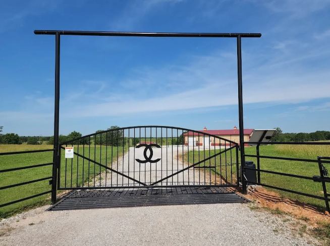 A black metal driveway gate with a central interlaced double-C emblem, standing on a gravel path under a blue sky.