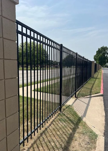 A black metal fence stretches into the distance, attached to a tan masonry pillar, alongside a sidewalk and grassy area.