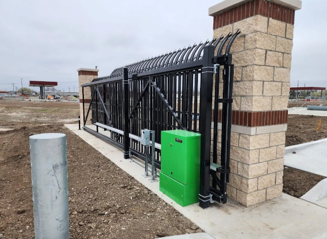 A black metal sliding security gate attached to a stone pillar, with a green control box, set in a dirt and concrete lot.