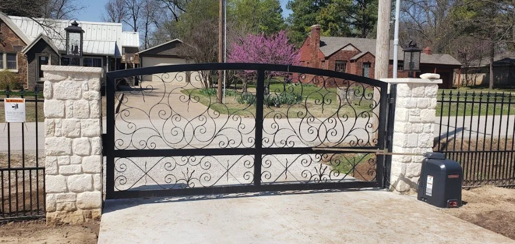 A black wrought-iron driveway gate sits between two light-colored stone pillars in a residential neighborhood.
