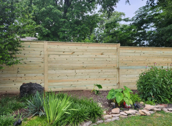 A horizontal wooden privacy fence stands in a backyard behind a garden bed with lush green plants and decorative rocks.