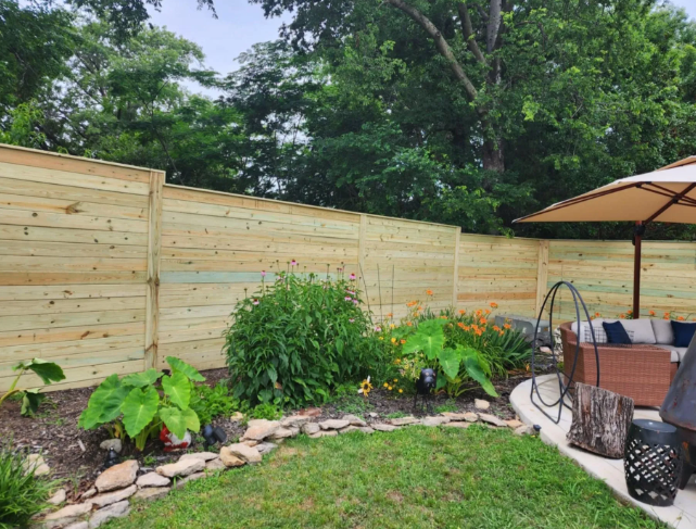 A new light-wood horizontal fence lines a lush backyard garden bed with green plants, stones, and a patio furniture set.