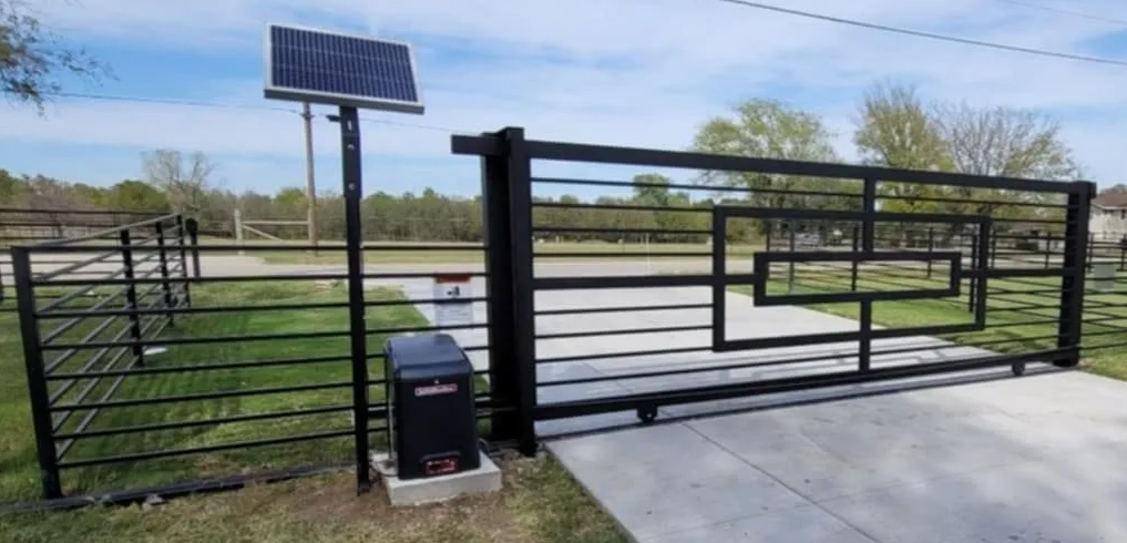 Black metal sliding driveway gate with a solar-powered motor and panel, set against a rural landscape.