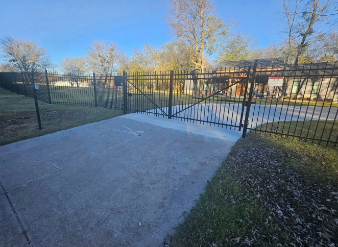 A driveway leading to a black wrought iron gate on a sunny day with trees in the background.