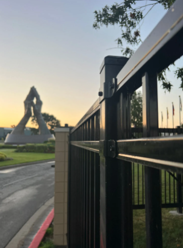 A black metal fence in the foreground with a large, abstract stone sculpture of hands held in prayer against a sunset.