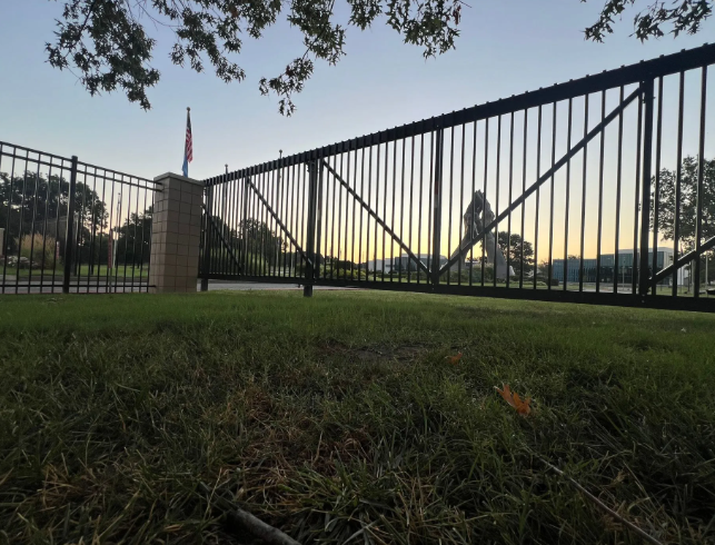 A low-angle view of a black metal fence at sunset, with a large, abstract metal sculpture visible in the background.