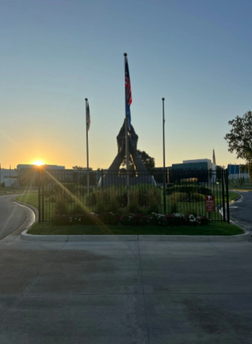 A sunrise silhouette of a tall monument with three flagpoles behind a black fence, set against a clear blue sky.