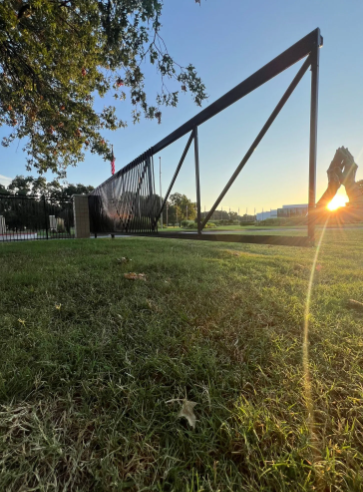A low-angle view of a triangular metal frame structure on a grassy lawn at sunset, with light flaring from the sun.