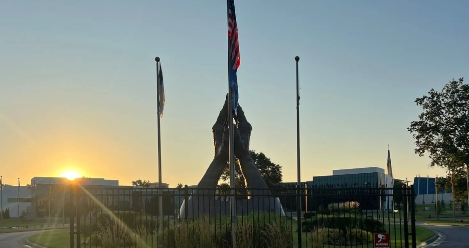 A monument of two praying hands stands at sunrise between three flagpoles, framed by a fence and nearby buildings.