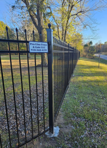 A black metal ornamental fence line runs across a grassy field under a clear blue sky, with a branded sign on a post.