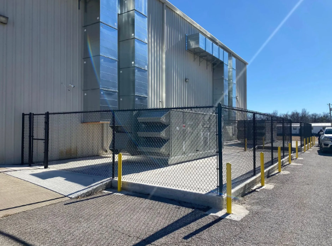 An industrial building with metal ventilation ducts and a large HVAC unit secured behind a black chain-link fence.