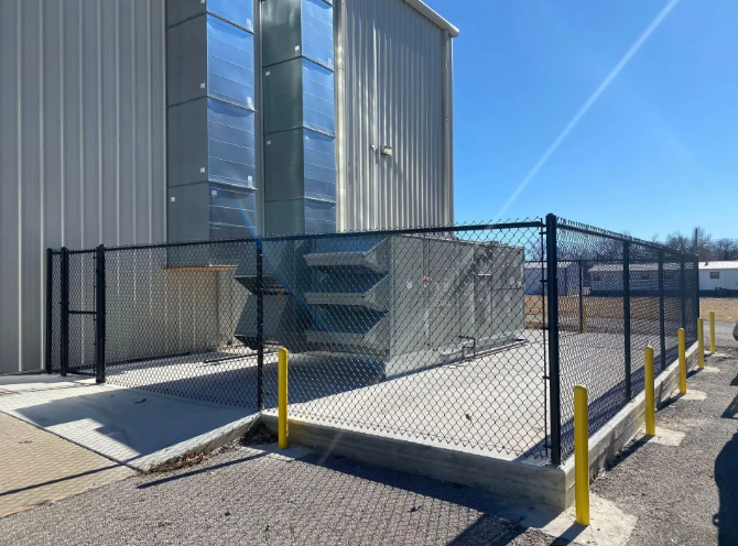 A metal building exterior with a large industrial HVAC unit inside a fenced-in concrete enclosure under a clear sky.