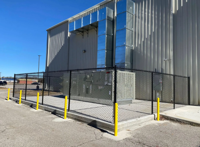An outdoor HVAC unit enclosed by a black chain-link fence on a concrete pad next to a metal-sided building.