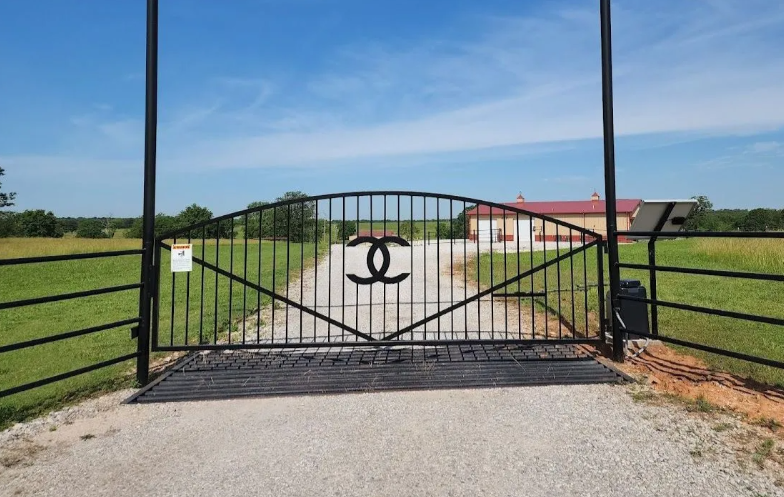A black metal gate featuring a central interlocking C logo, set on a gravel driveway leading to a building in a rural field.