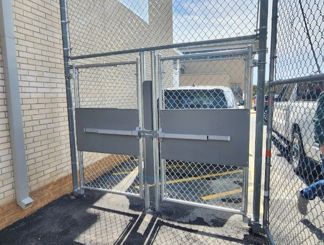 A chain-link gate with double doors and horizontal gray push-bar emergency exit hardware mounted on an exterior brick wall.