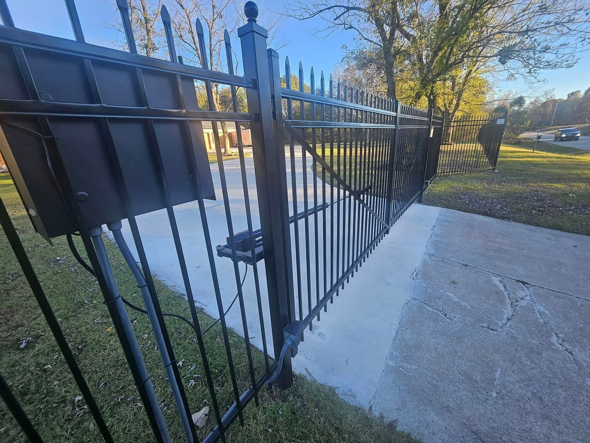 A black metal gate with an attached control box, partially open over a concrete driveway near a grassy area.