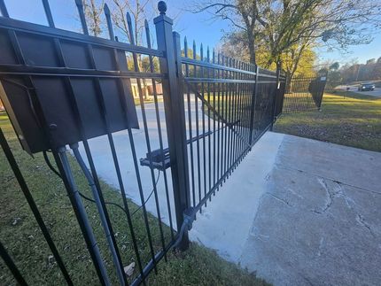 A black metal gate with an attached control box, partially open over a concrete driveway near a grassy area.