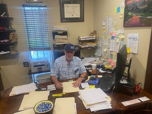 Man sitting at a cluttered office desk, writing papers in a small room with notes and shelves behind him.