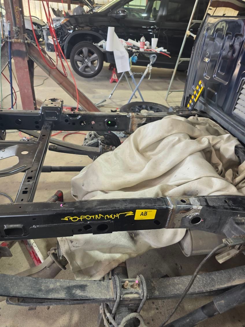 Destroyed vehicle frame with a beige tarp draped over it in a garage or repair shop; car visible in background