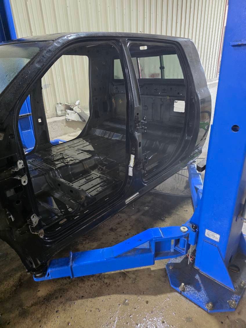 Black vehicle body shell lifted by a blue forklift in a workshop, with doors missing and interior exposed