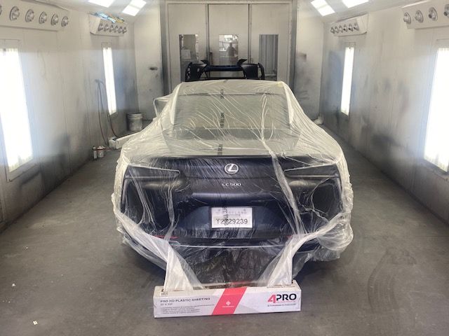 Black car wrapped in plastic inside a white spray booth, with a paint sample board in front