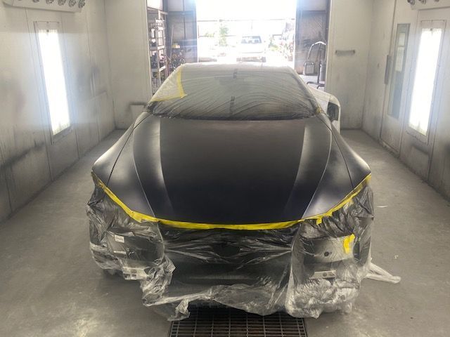 Black car in a spray booth, front covered with plastic and yellow tape during painting.