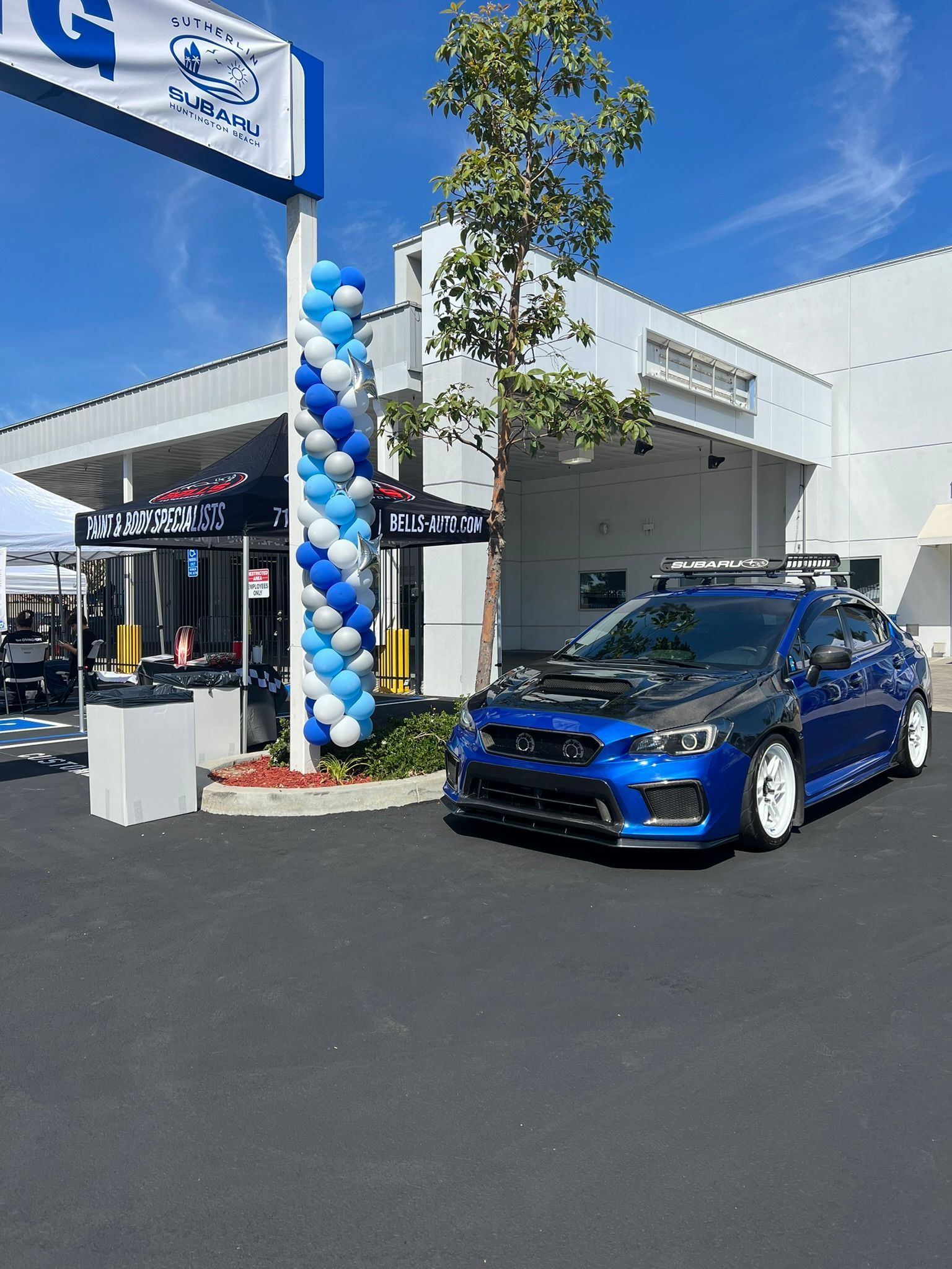 Blue sports car parked outside a dealership with blue-and-white balloons under a clear sky