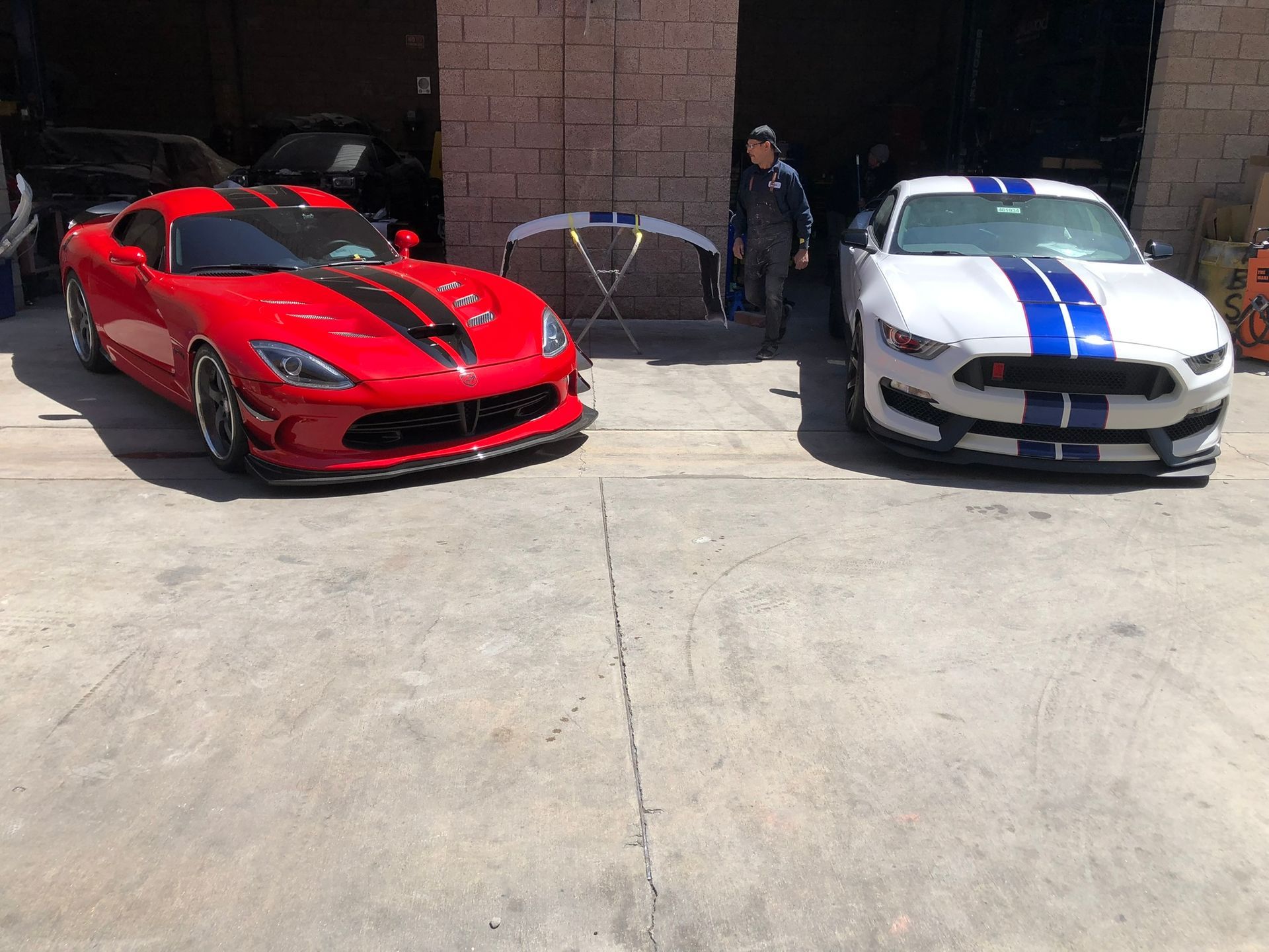 Two sports cars, one red and one white with blue stripes, parked in front of a garage with a person standing between them