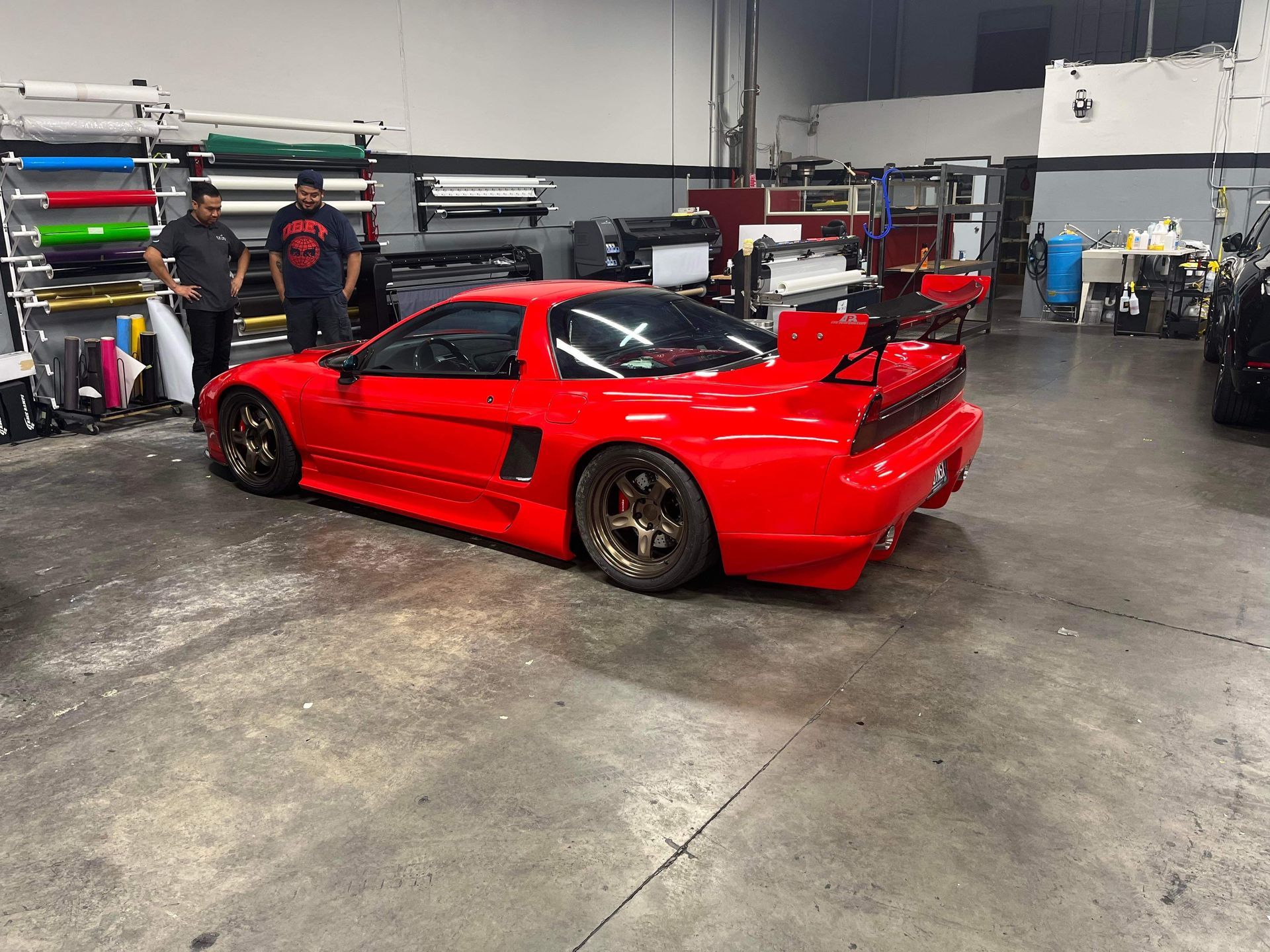 Red sports car with a large rear wing in an auto shop garage with two people nearby.