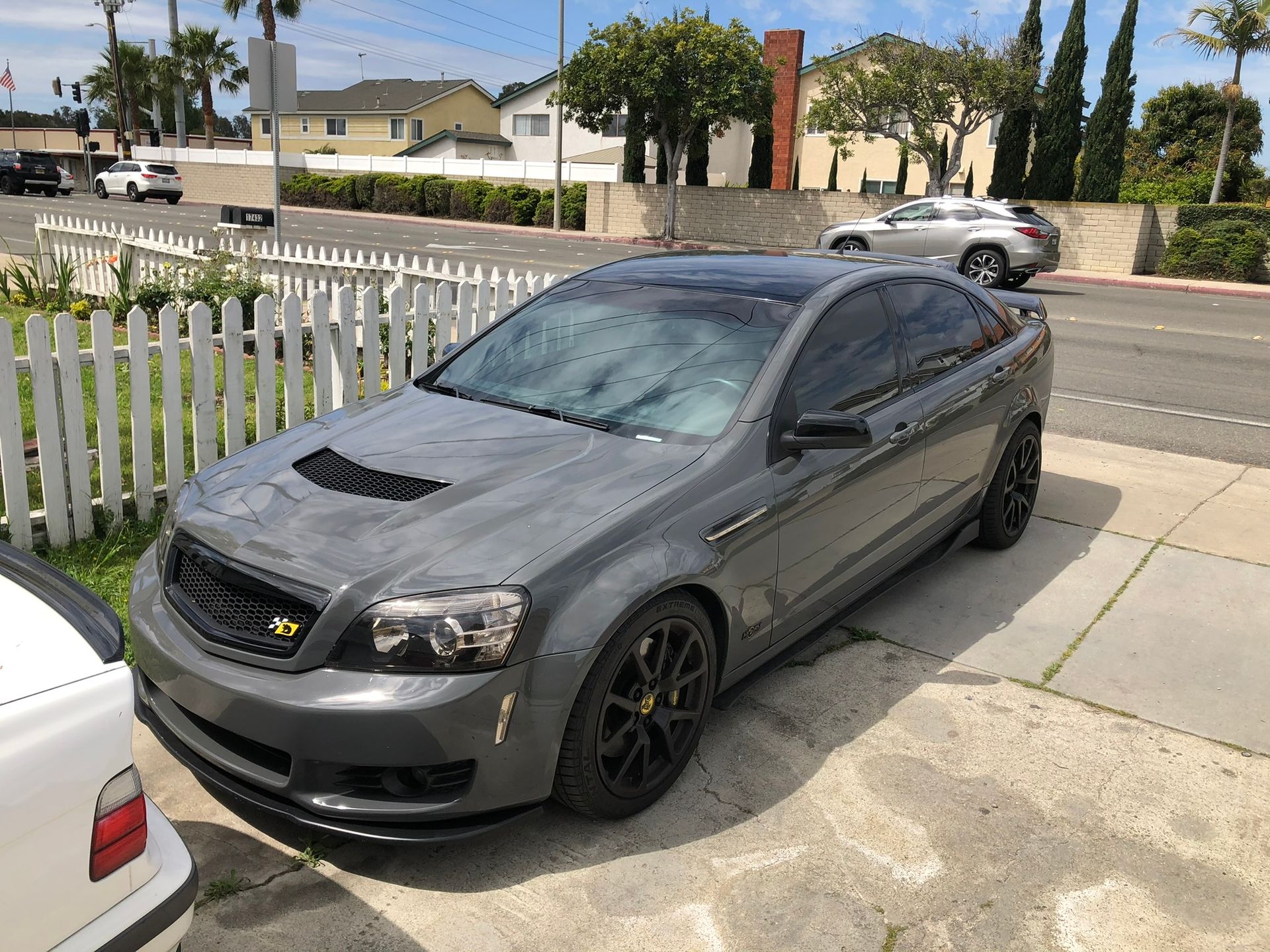 Dark gray sedan parked in a driveway beside a white fence on a sunny residential street.