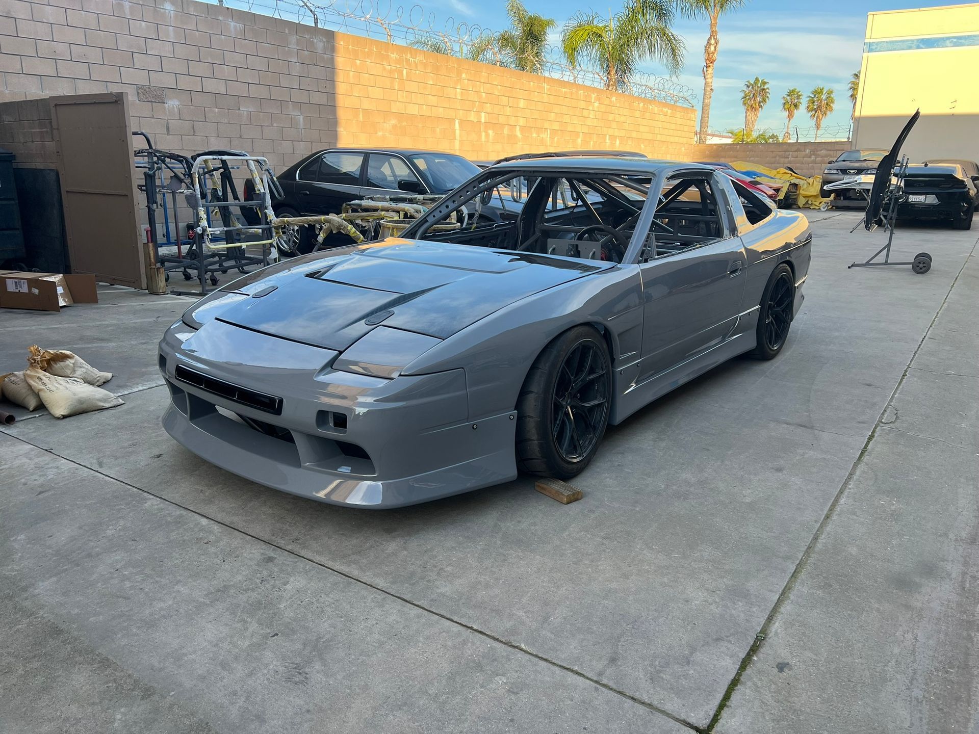 Gray sports car parked in an outdoor lot with another car and a brick wall in the background