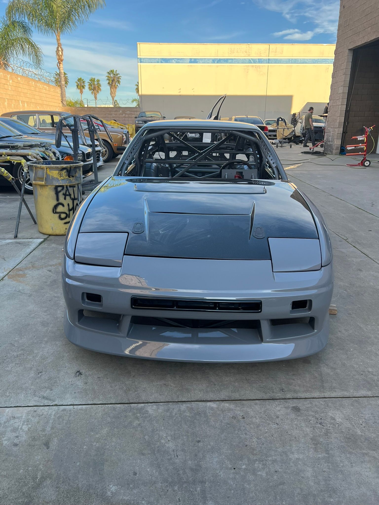 Gray sports car in a repair yard, front view with hood open and missing headlights.