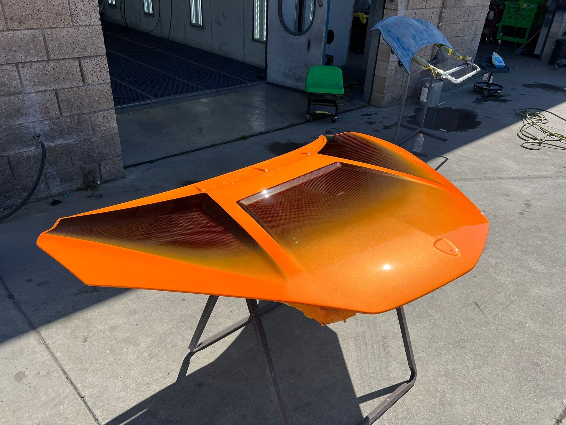 Bright orange sports car body shell on a stand outdoors near a garage.