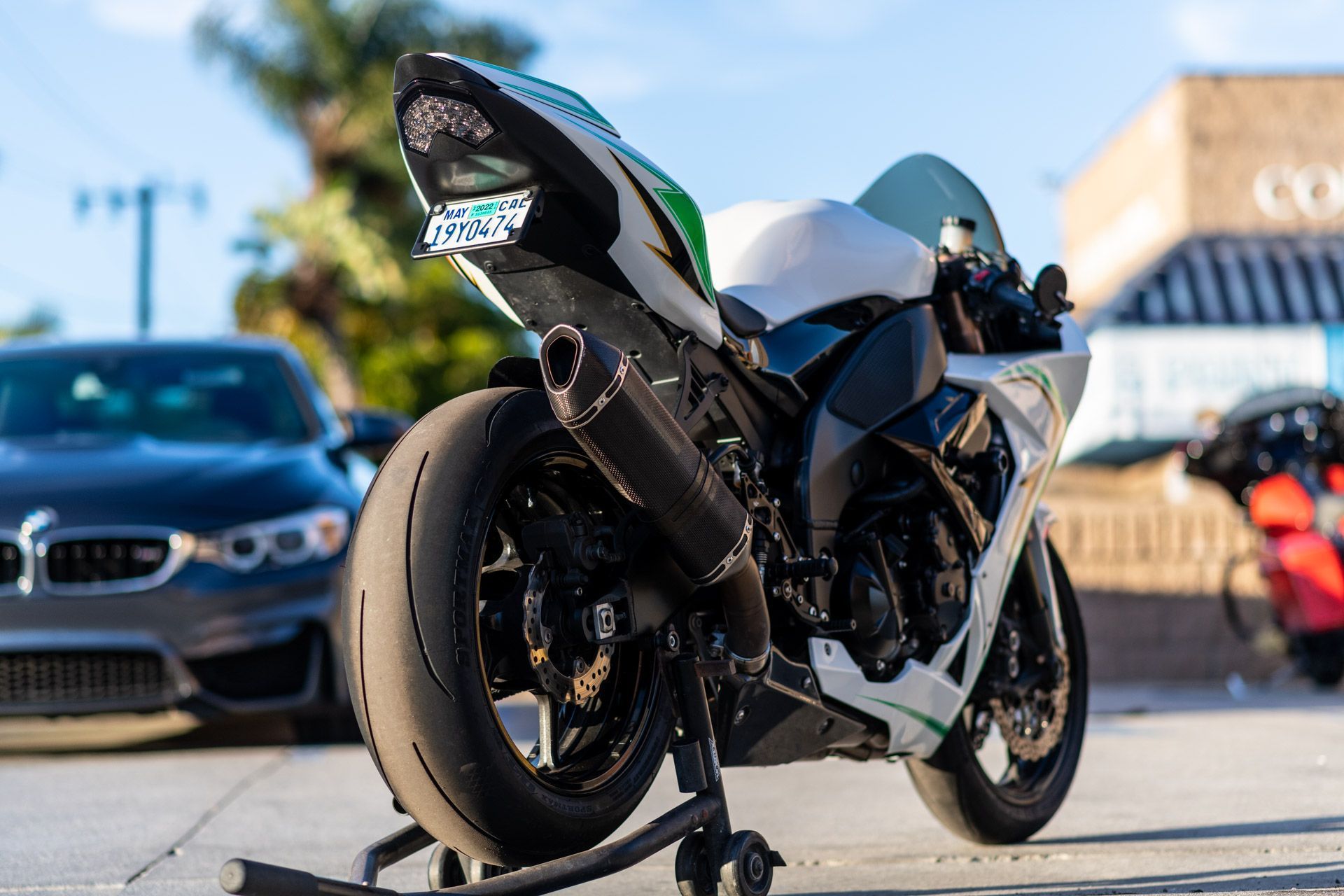 White sport motorcycle parked on a stand at a sunny street-side car meet, with cars blurred in the background