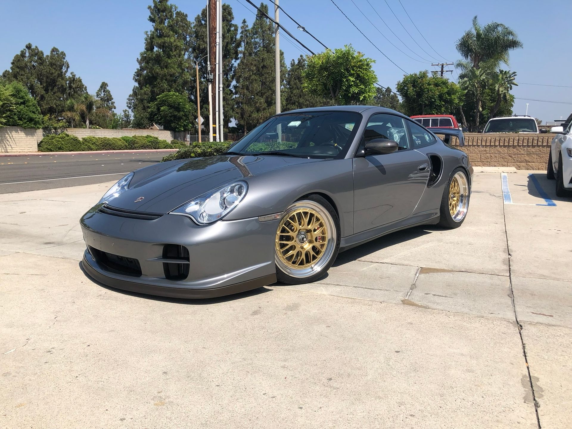 Gray sports car with gold wheels parked on a sunlit concrete lot.