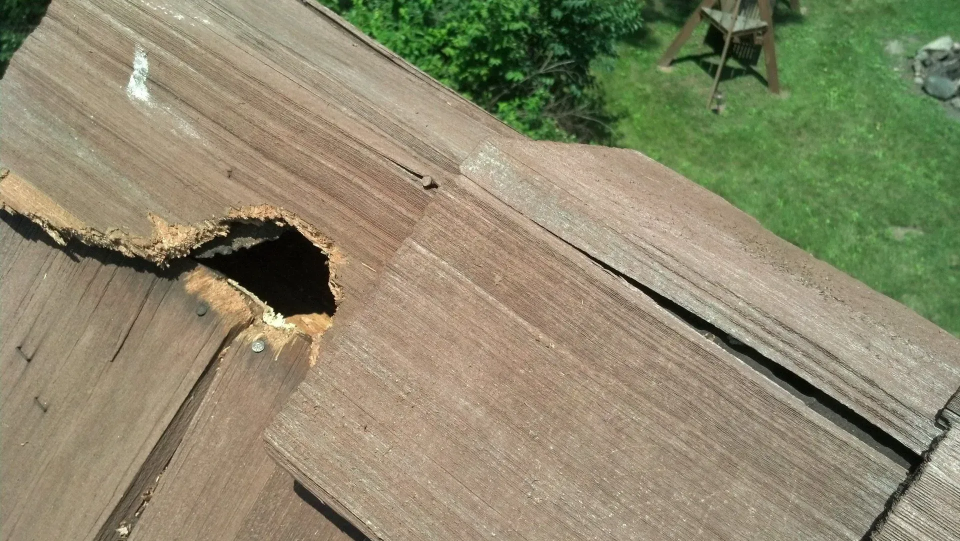 Close-up of a damaged brown shingle roof with a large hole, set outdoors.