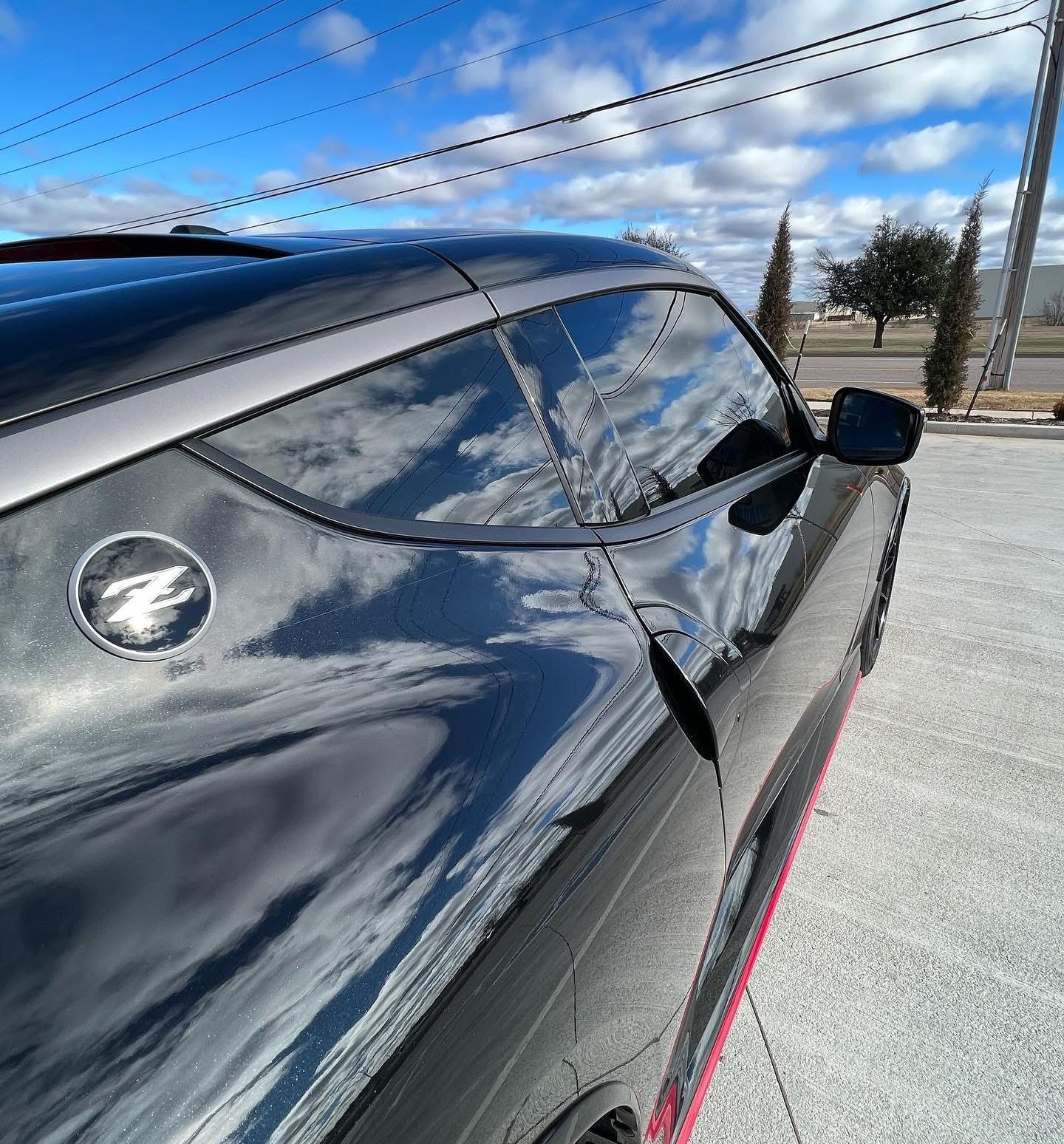 Black Nissan Z sports car, reflecting clouds, with a gray roof and Z emblem.