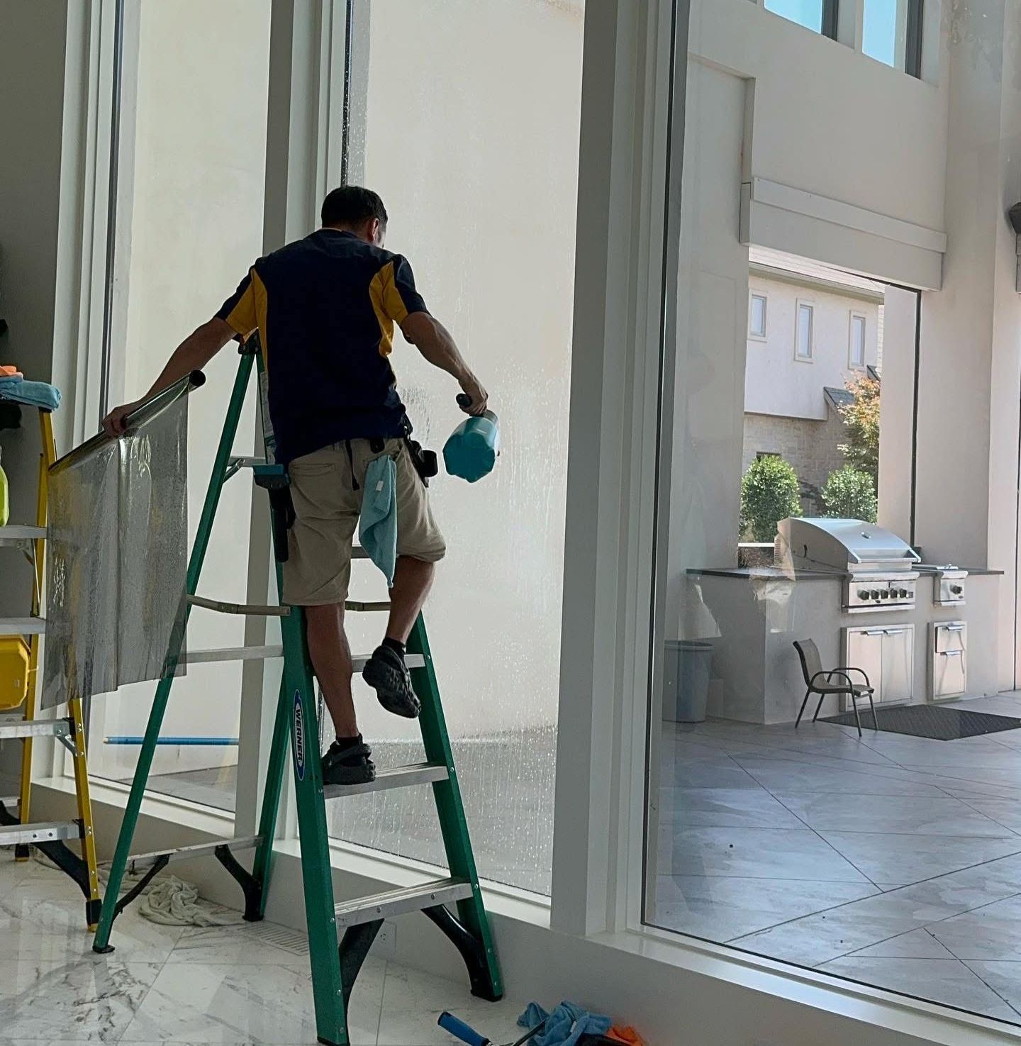 Man on ladder cleaning a large window; he sprays it in a bright indoor space with an outdoor patio visible.