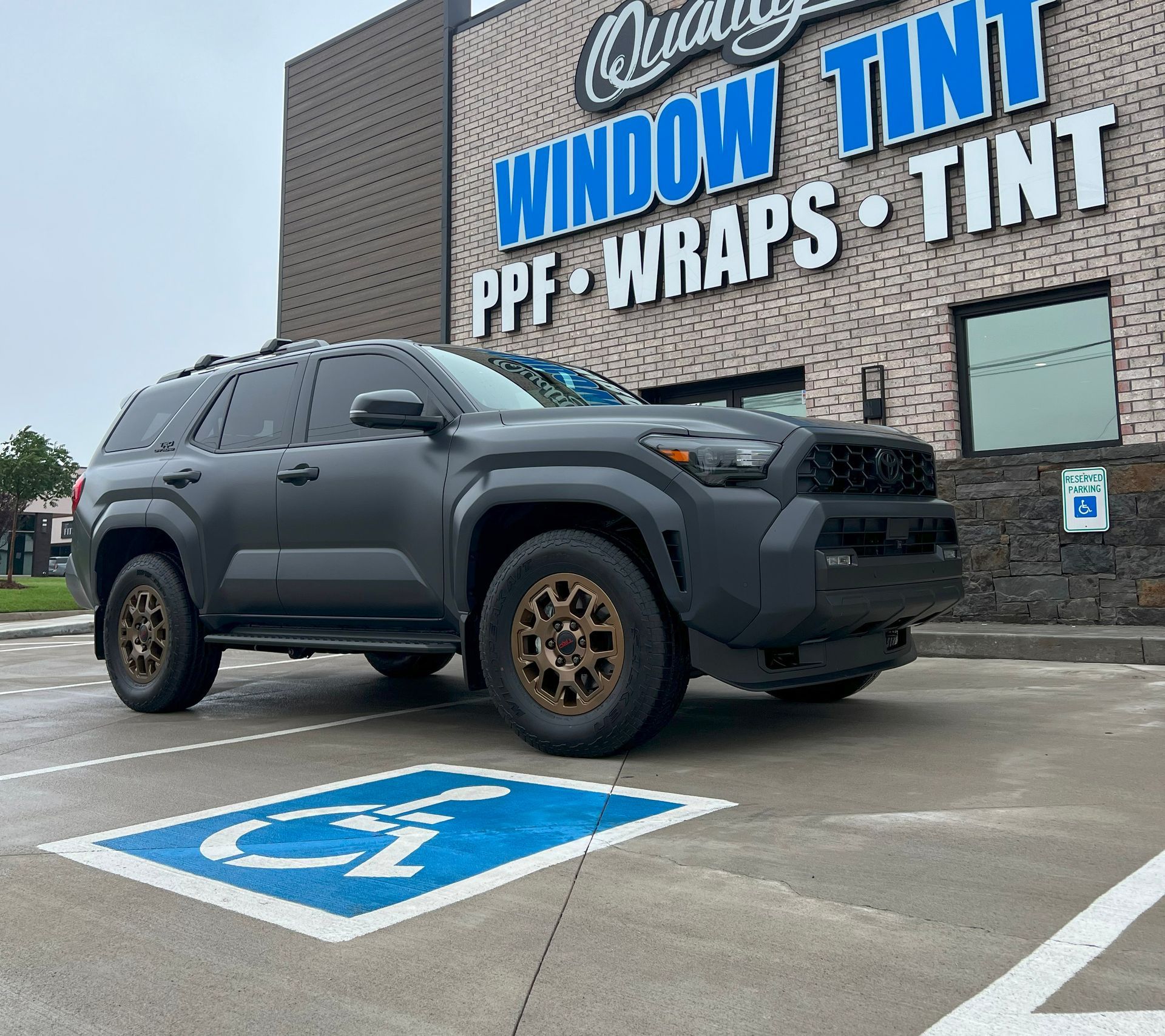 Dark grey Toyota Sequoia SUV with bronze wheels parked in a handicap spot in front of a window tint shop.