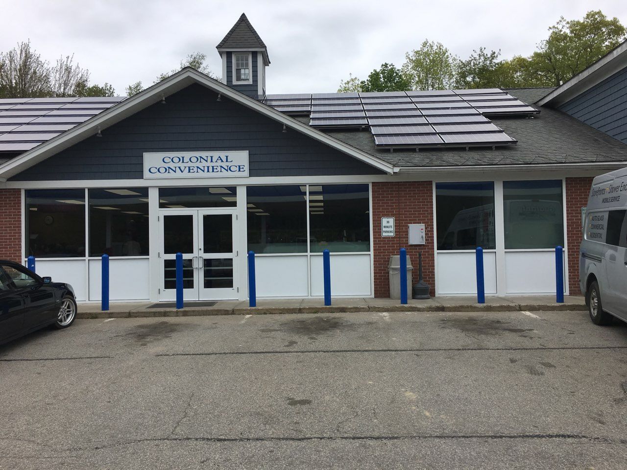 Colonial Convenience store exterior with solar panels on roof. Brick and white building with dark blue trim, parked cars.