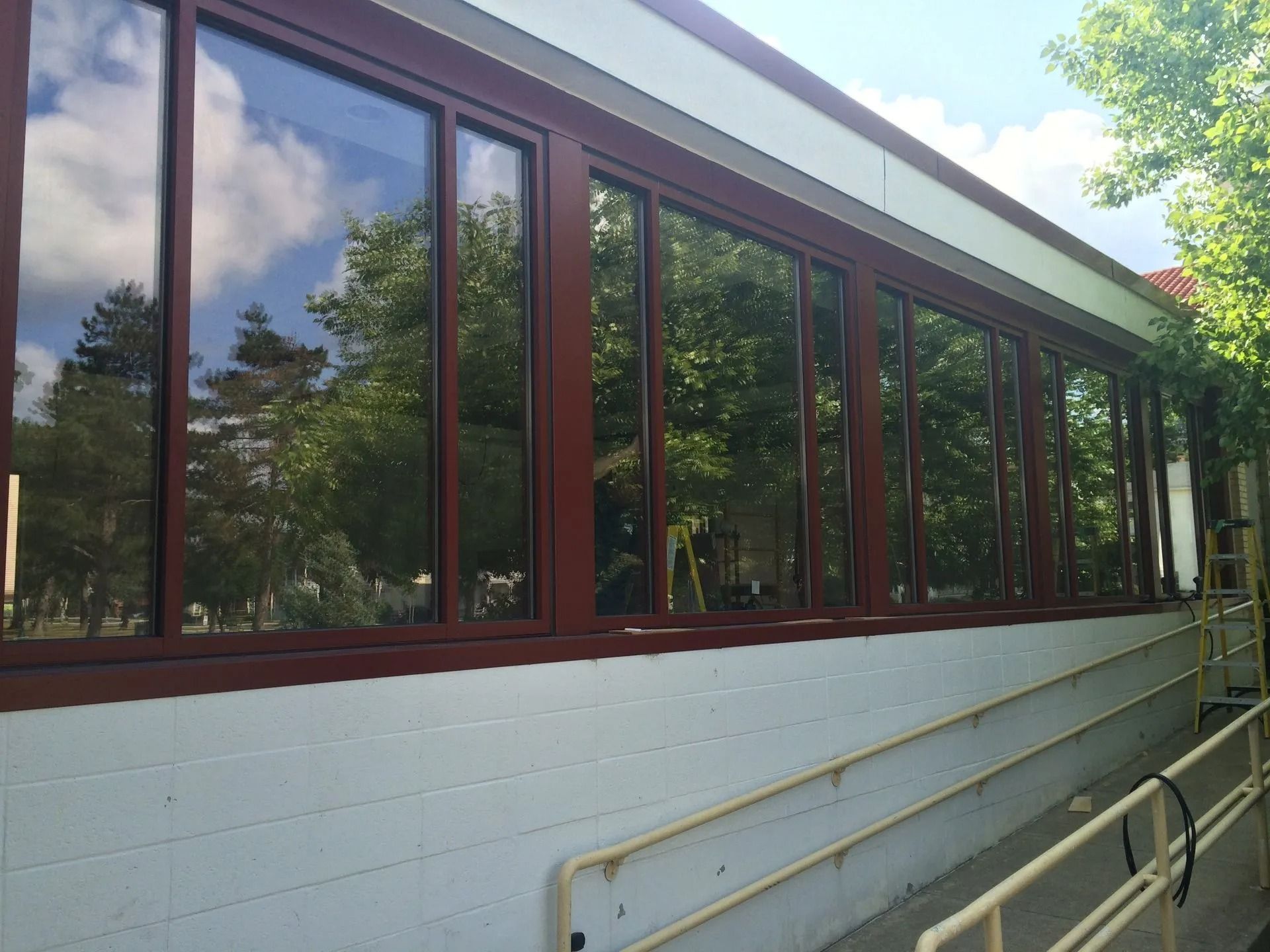 Long row of windows with brown frames on a white building, reflecting trees and sky. A ramp is visible.