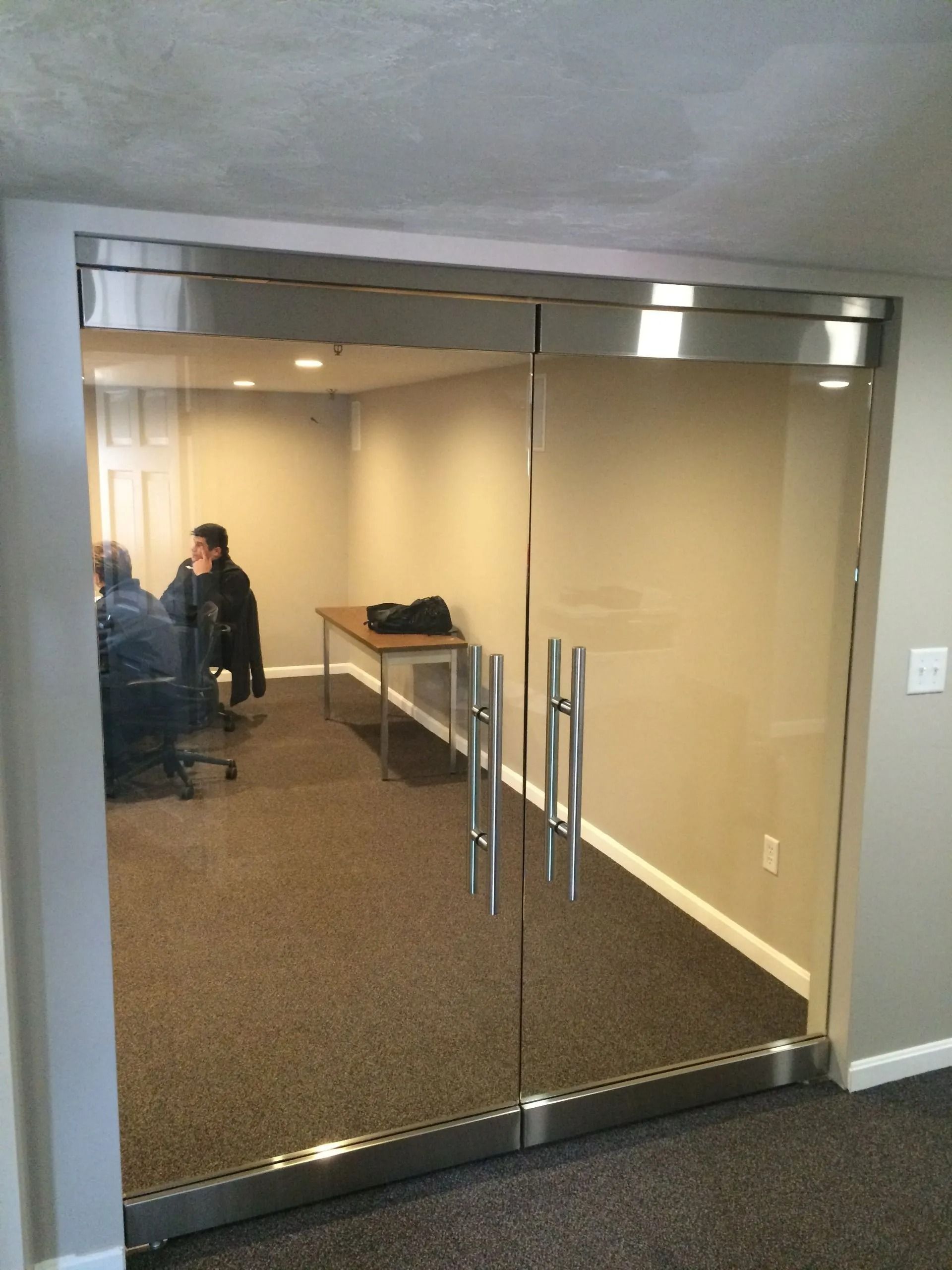 Glass door leads to an office with people, table, and dark carpet. Chrome door handles and trim.