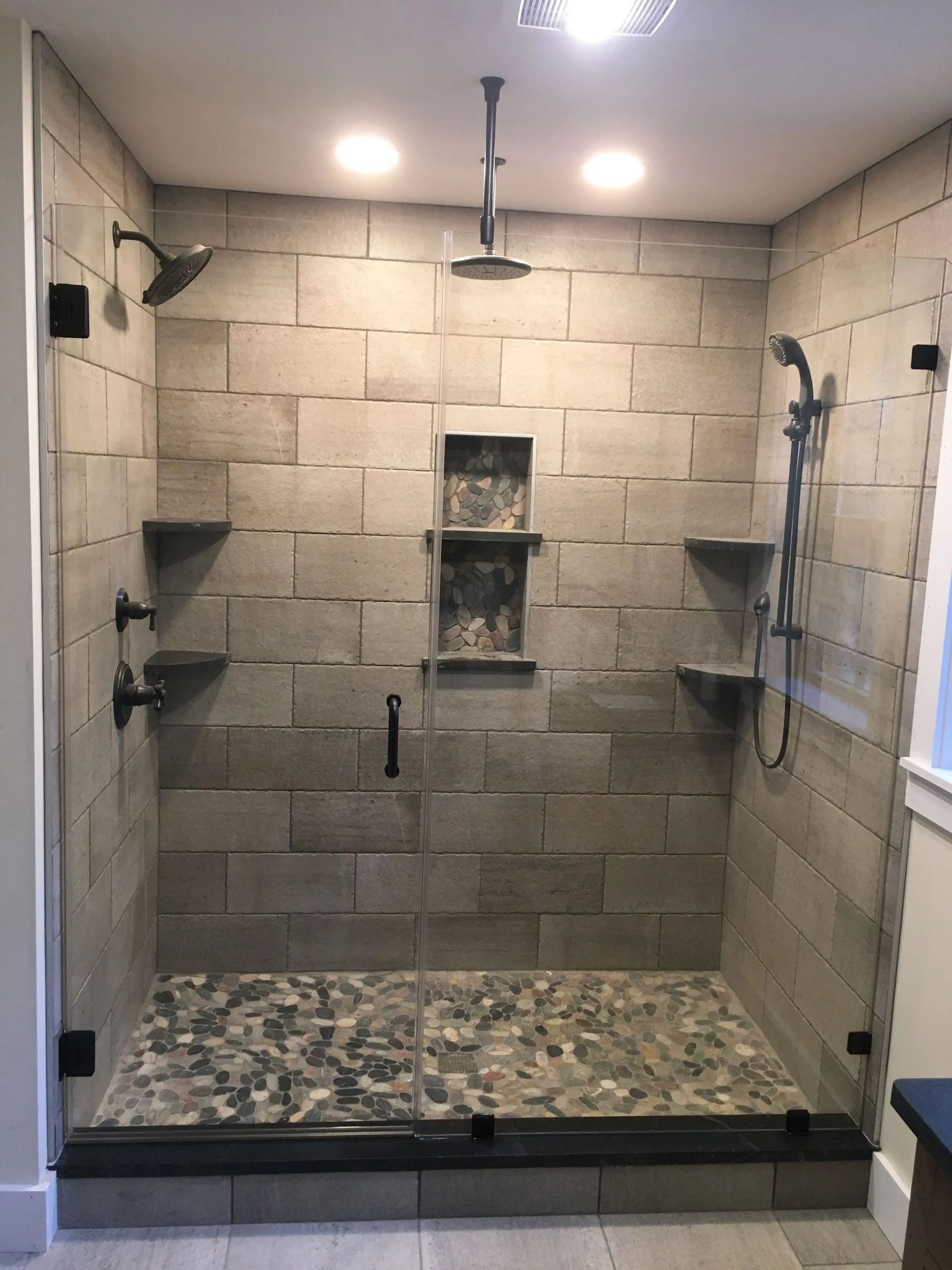 Modern shower with stone tile walls, pebble floor, glass doors, multiple showerheads, and built-in shelves.