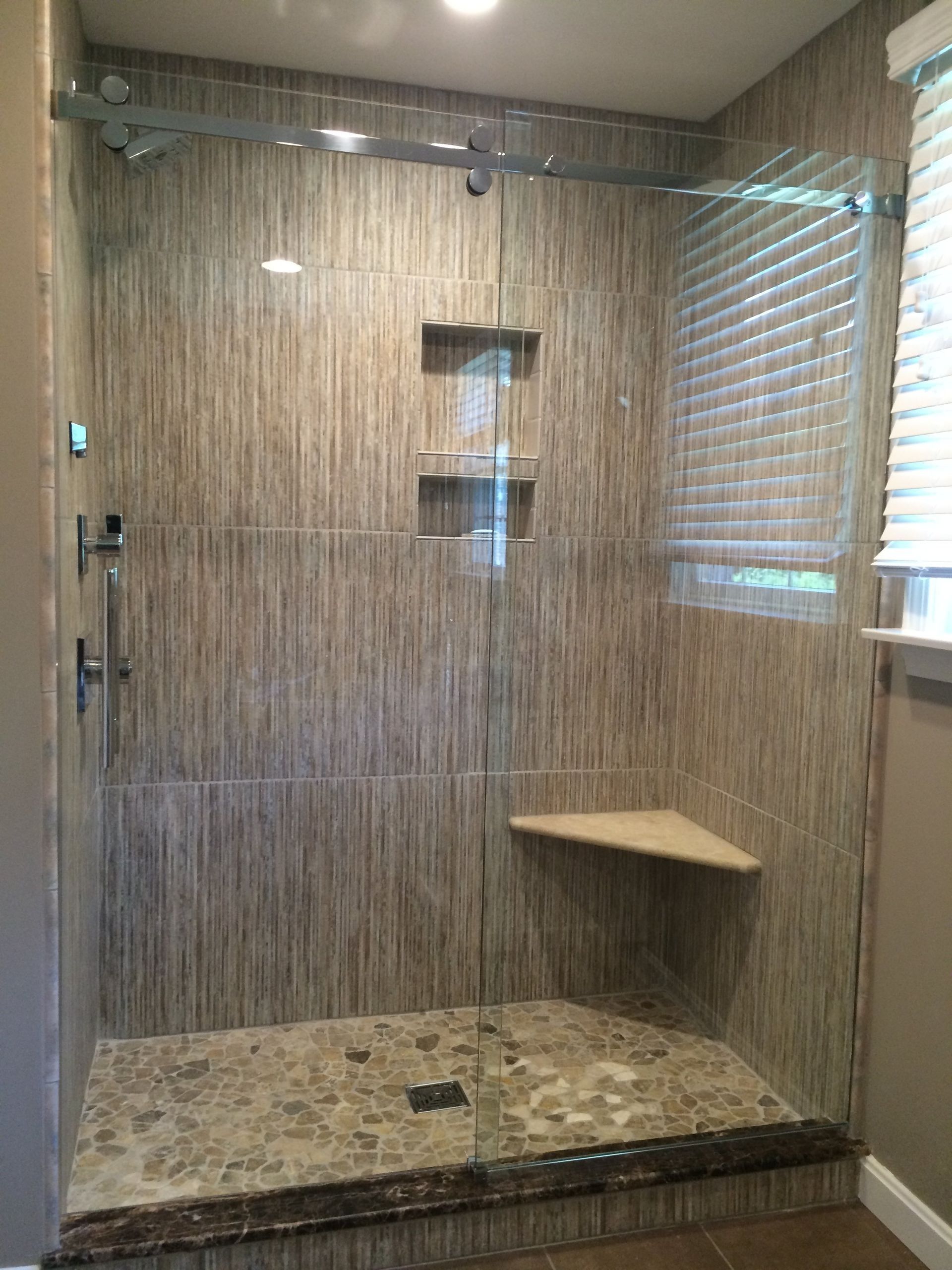 Modern shower with tan textured tile walls, clear glass enclosure, built-in shelves, and a corner seat.
