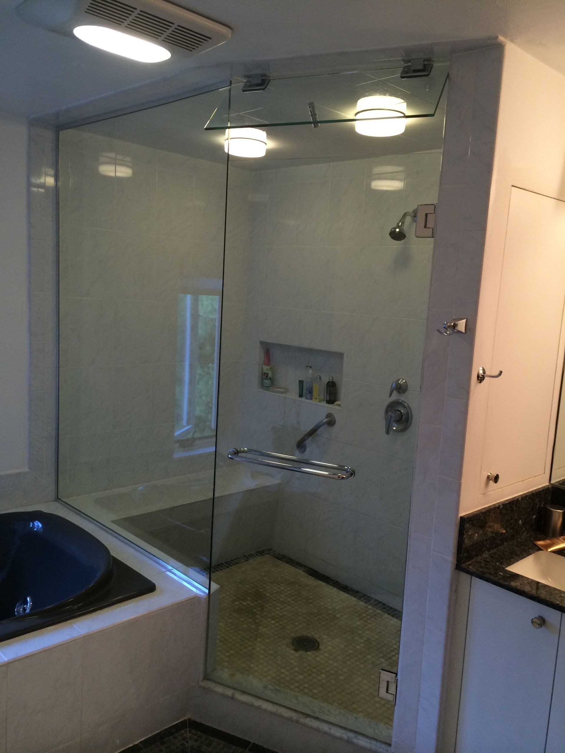 Glass-enclosed shower stall with a tub. White walls, chrome fixtures. Three lights.